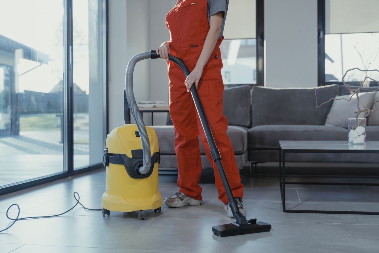 services-02 A professional cleaner in red coveralls vacuuming a modern living room with a yellow vacuum cleaner.